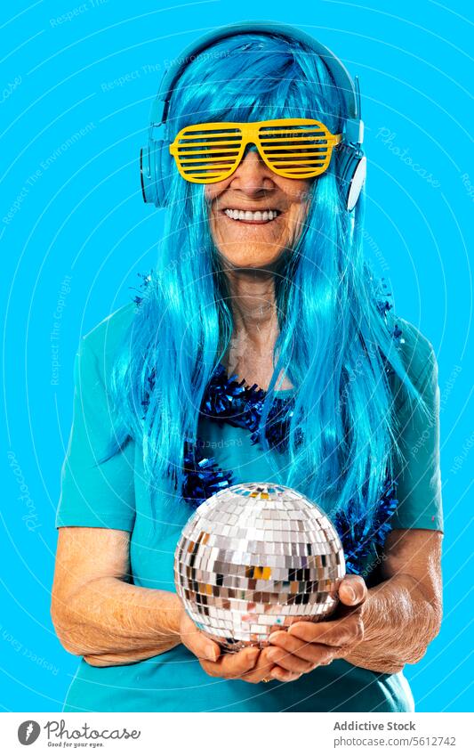 Cheerful aged woman in blue wig standing with disco ball and shutter glasses listening to music on headphones against blue background confident party fashion