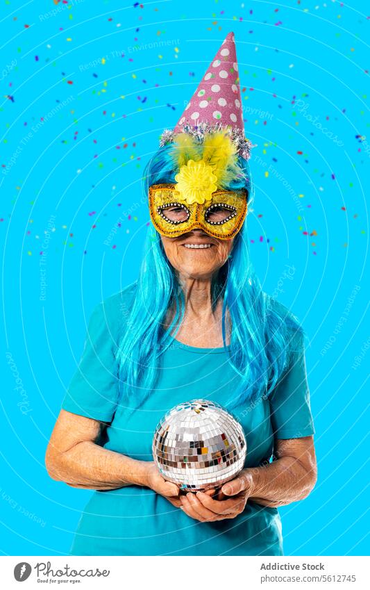Happy elderly woman wearing carnival mask, blue wig and party hat and holding a disco ball while standing on blue background in studio celebrate confetti