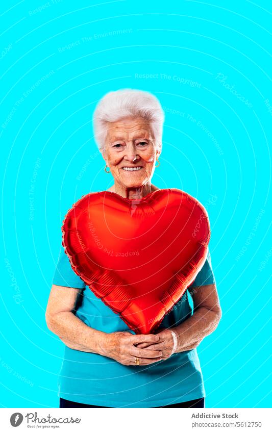 Smiling senior woman holding heart shaped balloon and looking at camera against blue background love valentine saint valentine day romantic studio shot retire