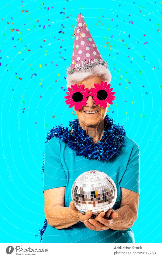 Smiling senior lady wearing party cap, funny sunglasses and tinsel around neck holding disco ball on blue background woman new year celebrate having fun