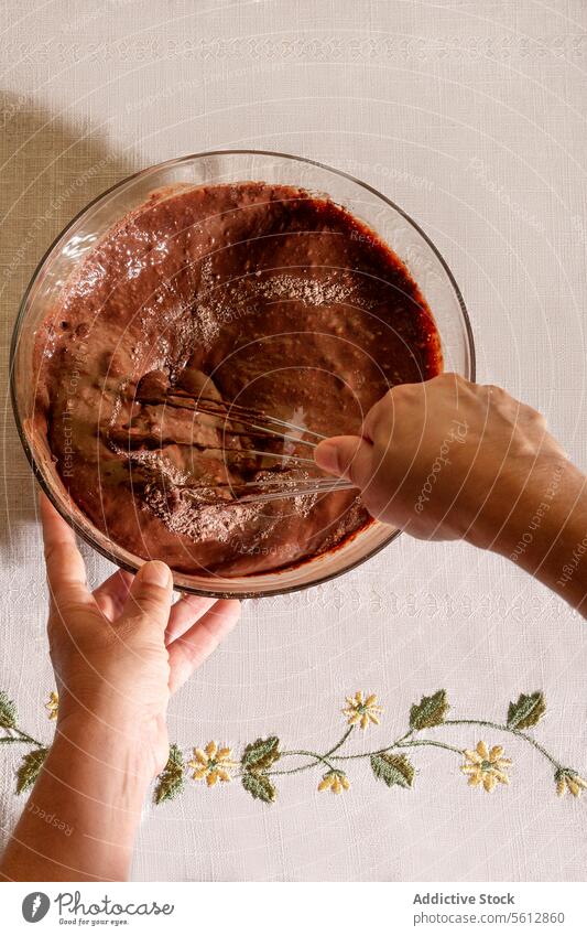 Top view of unrecognizable person Whisking Chocolate Batter in a Glass Bowl Over Floral Cloth hand whisk chocolate batter glass bowl floral cloth embroidered