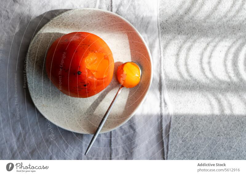 Gleaming Persimmon on Plate with Shadow Play persimmon plate spoon shadow ripe fruit dessert ceramic vibrant dramatic curve gleaming seasonal tabletop fabric