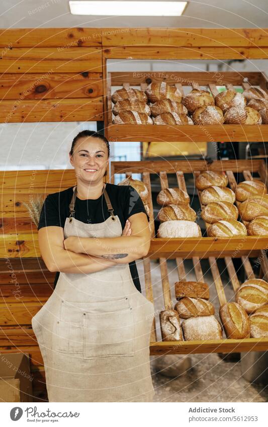 Happy saleswoman standing by bread in bakery shop seller shelf loaf confident smile arms crossed apron fresh baked food wooden positive manufacture production