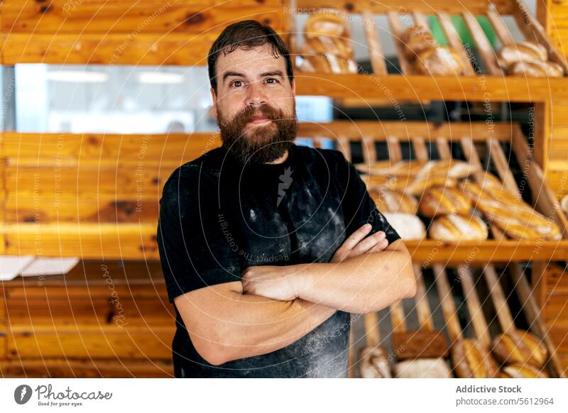 Calm salesman standing by bread in bakery shop confident shelf arms crossed store blur seller loaf beard casual attire fresh calm baked food wooden positive