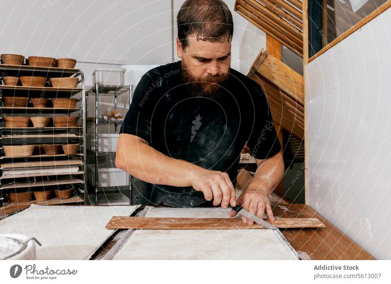 Chef cutting pastry dough in bakery wooden tool knife focus serious concentrate table bakehouse uncooked utensil mold casual attire standing holding man beard