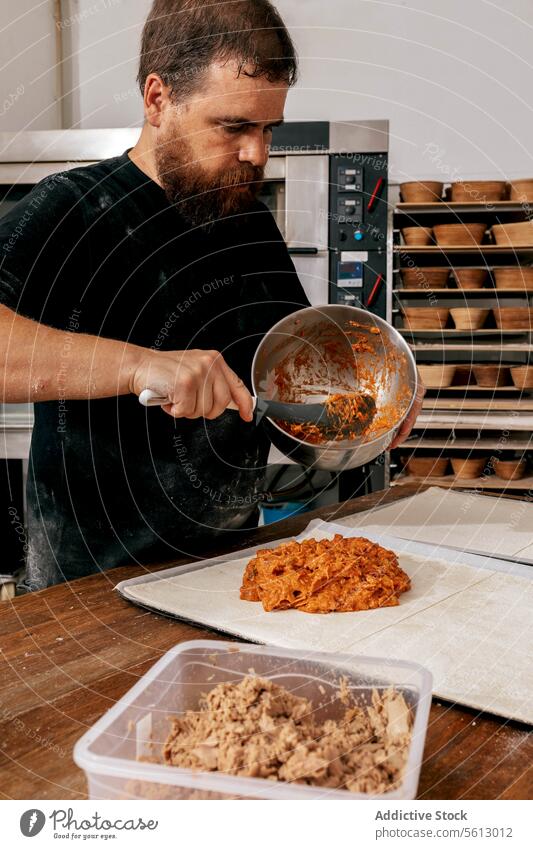 Male chef making pastries on table in bakehouse baker pastry stuffing dough bakery kitchen serious concentrate casual attire bowl spoon container utensil man