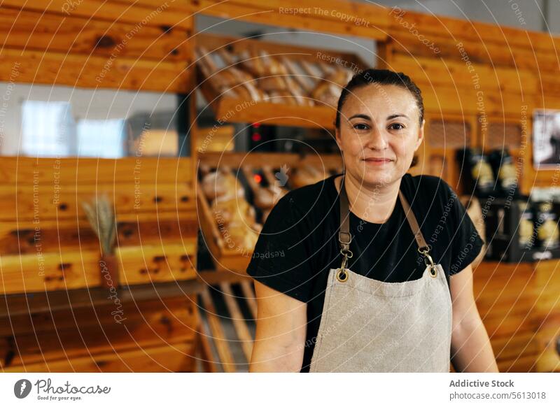Happy saleswoman standing by bread in bakery shop seller shelf loaf confident smile apron fresh baked food wooden counter positive manufacture production