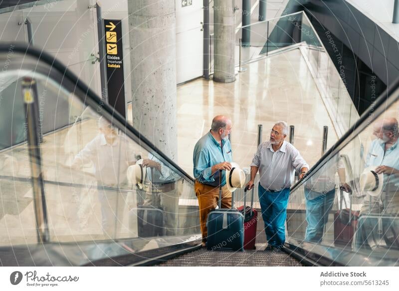 Friends with baggage on escalator at airport luggage talk traveler men terminal high angle stand moving stairs suitcase hat transit friends senior vacation