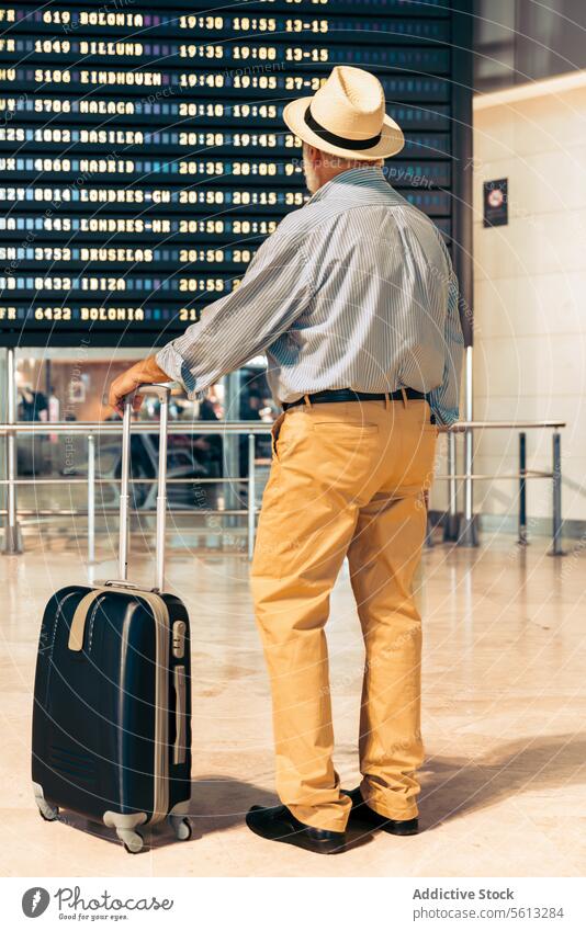 Man with luggage looking at timetable at airport senior man departure board back view suitcase vacation travel retired lifestyle leisure hat full body passenger