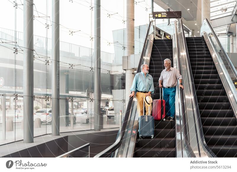 Friends with baggage on escalator at airport luggage talk traveler men terminal low angle stand moving stairs suitcase hat transit friends senior together