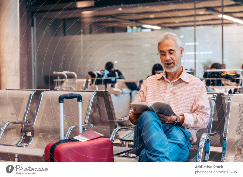 Aged man reading book at airport passenger chair beard eyeglass casual attire sit wait lounge hobby learn focus serious concentrate educate senior departure