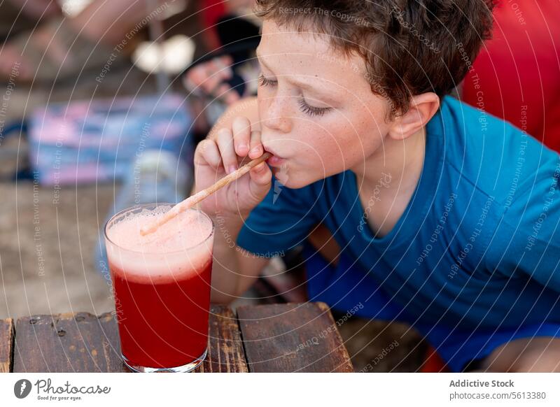 High angle view of little boy in casual clothes drinking fresh watermelon juice in glass with straw while sitting at wooden table in restaurant innocent cafe