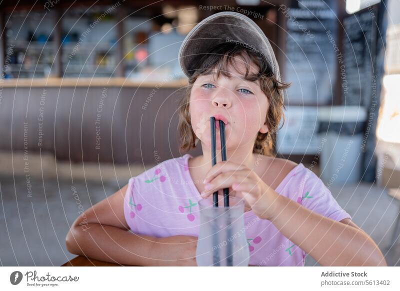 Child with straw in mouth sitting in cafe girl cap cute caucasian drinking contemplation summer fruit vacation thirsty pensive restaurant person lifestyle kid