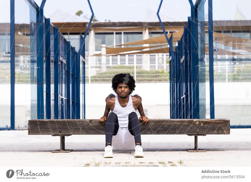 Full body of confident serious African American male athlete balancing body on wooden bench and doing squatting exercise while looking at camera in city man
