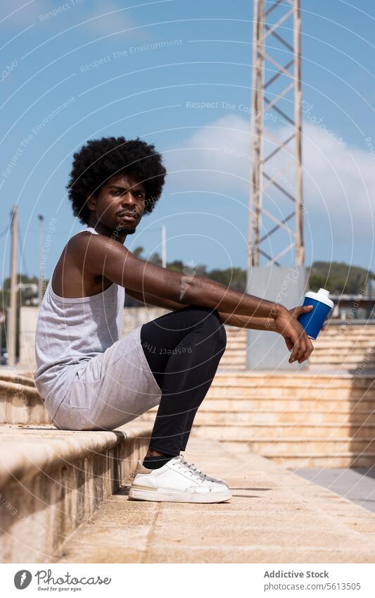 Side view of African American tired male athlete in sportswear sitting on staircase and with bottle water while having break from training on sunny day man