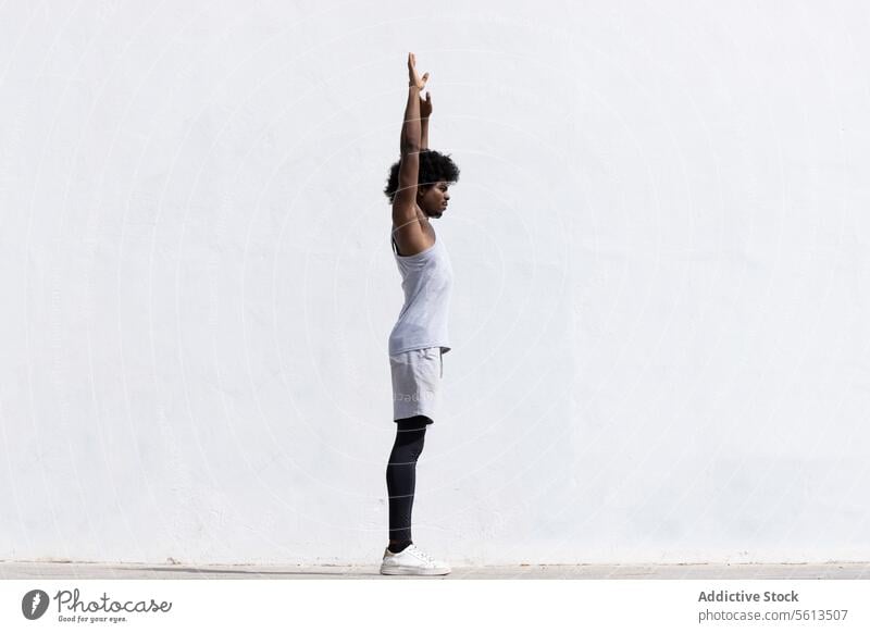 Full body side view of confident African American male athlete in activewear with concentration stretching arms while standing looking away against white background