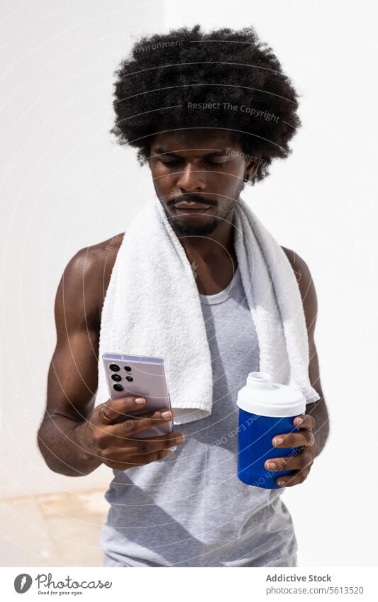 African american young sportsman with afro hair and towel on neck looking at screen while browsing over mobile phone and holding bottle of water against white wall