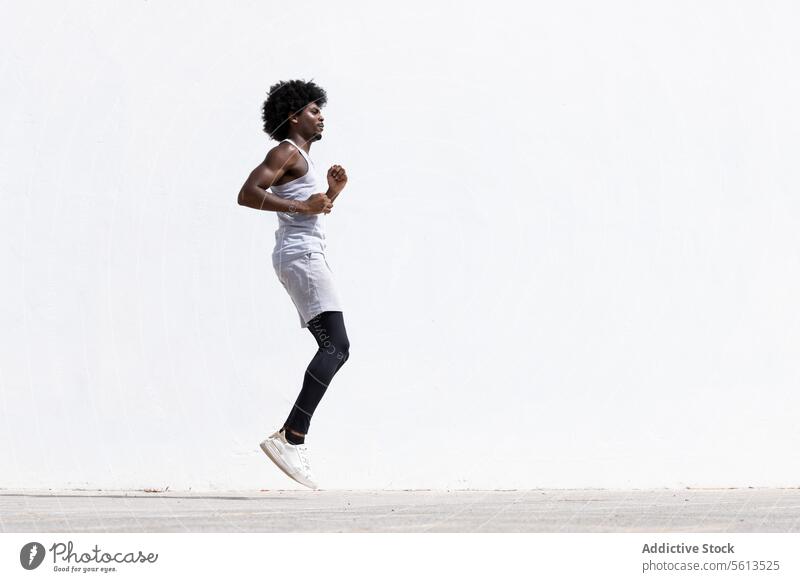 Side view of determined young active African American male athlete in sportswear jumping on street against white wall during fitness workout man exercise