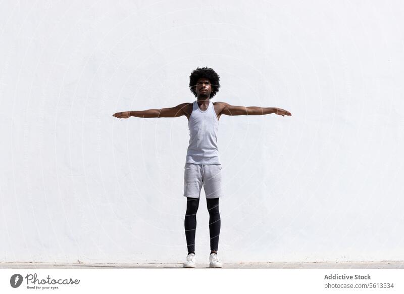 Full body of confident African American male athlete in activewear with concentration stretching arms while standing looking away against white background man