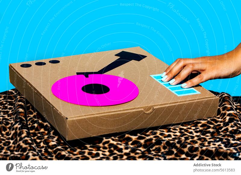 Crop hand of anonymous woman playing antique turntable made with cardboard box on yellow table over animal print textile print on blue background using party