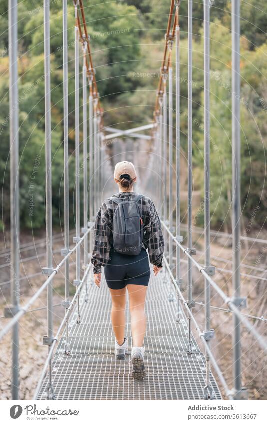 Back view of anonymous female hiker in shorts and backpack walking on narrow footbridge while trekking in forest during weekend holiday woman leg shoes explorer