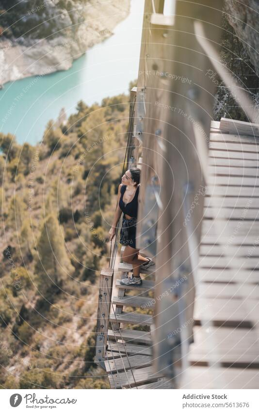 High angle full body of young female traveler looking away and admiring nature while standing on narrow wooden steps of mountain during weekend trekking admire