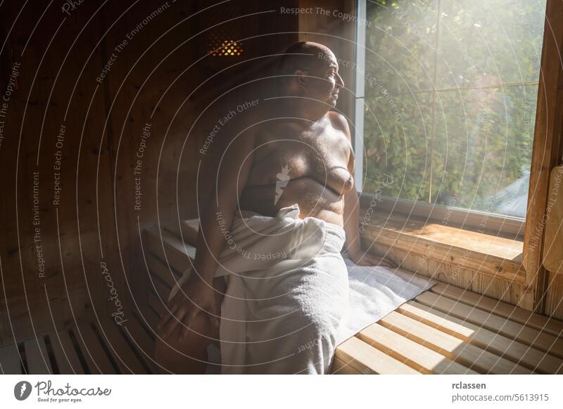 Man sitting in a sunlit finnish sauna, looking outside the window, wrapped in a towel wellness spa resort hotel sweat finland man in sauna smiling relaxation