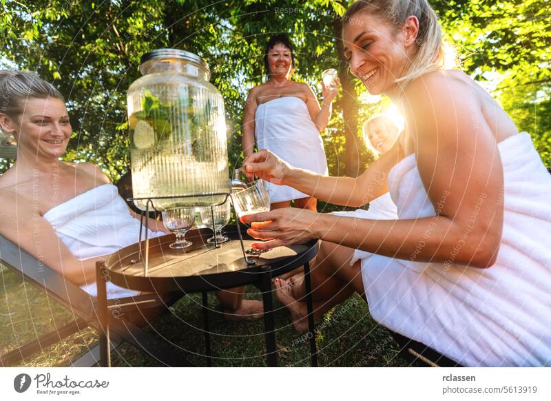 Women in spa towels serving infused water with limes from a dispenser in a garden setting after finnish sauna. lemon women beverage dispenser relaxation