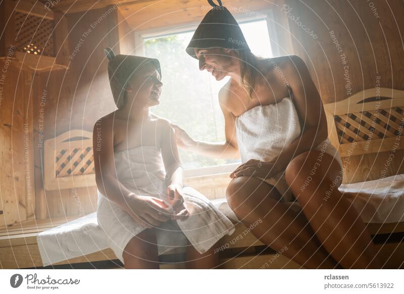 Mother and child smiling at each other in a finnish sauna, both wearing felt hats bathrobe steam spa wellness hotel calm daughter family woman relaxation