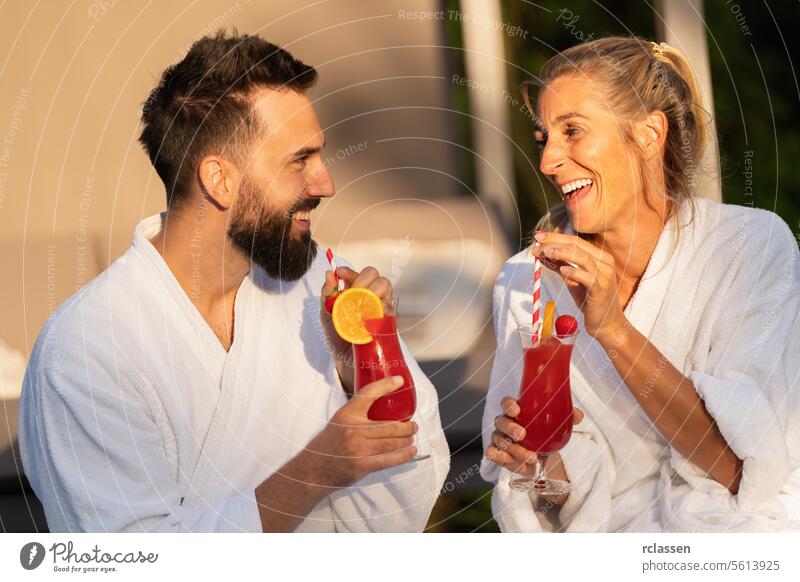 Two people in white robes laughing and drinking cocktails in the sunlight at a wellness hotel sipping bathrobe spa wellness resort couple red cocktails joyful