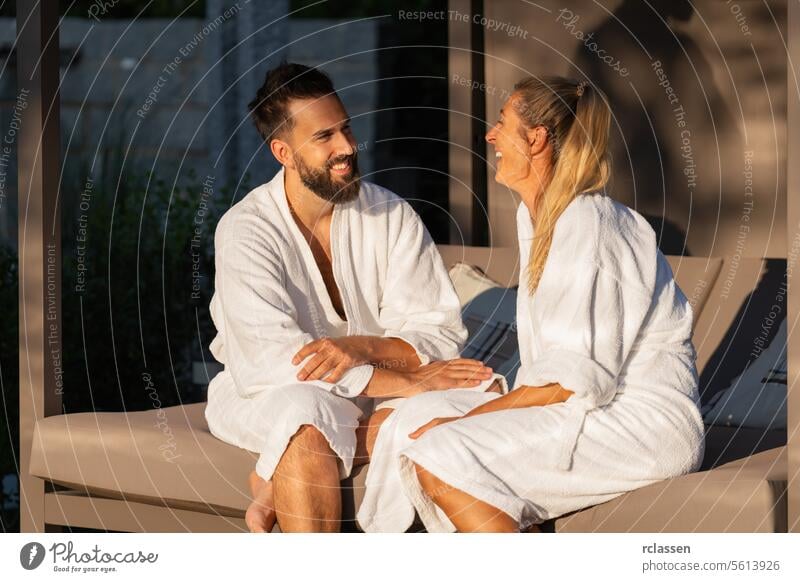happy couple in white bathrobes sitting and talking on an outdoor bench lounger in sunlight at spa wellness hotel smiling white robes relaxation happiness