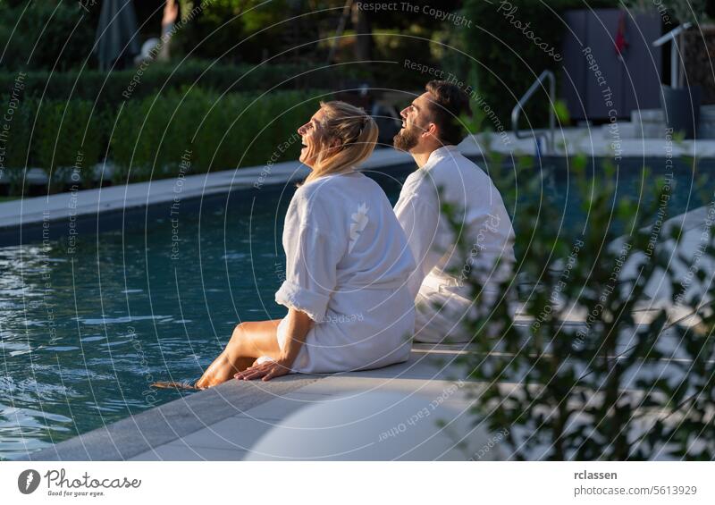 Couple in white robes seated by a pool looking up and laughing a day in a spa hotel wellness spa resort couple relaxation leisure happiness bonding joy