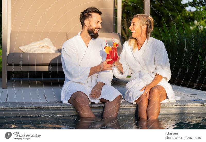 Two people in white bathrobes laughing and toasting with red drinks by a pool at sunset at a spa hotel relaxation cocktails friends laughter clinking glasses
