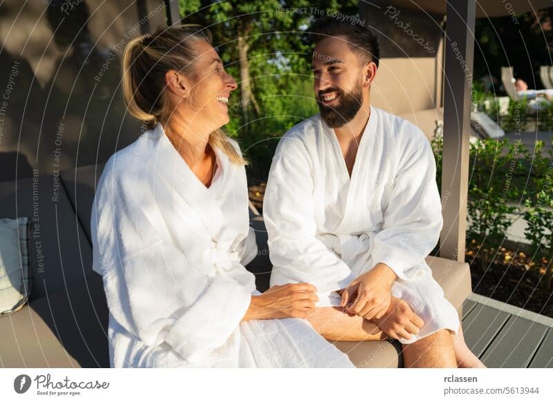 smiling couple in white bathrobes enjoying a sunny day outdoors on a couple's lounger at a spa hotel love laughing together resort wellness spa relaxation