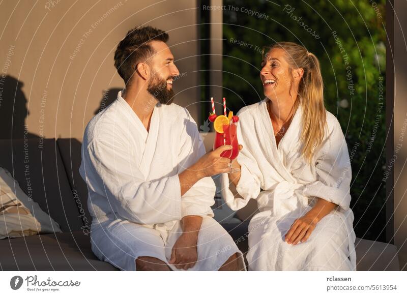 Smiling couple in white spa robes clinking cocktails in sunny ambiance at spa resort clinking glasses bathrobe wellness hotel spa wellness resort white robes