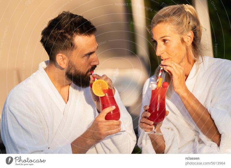 couple in white bathrobes drinking cocktails and looking at each other at a hotel sipping wellness spa wellness resort white robes red cocktails joyful