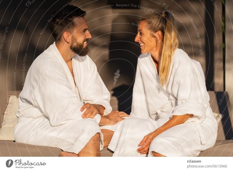 Happy couple in white robes talking and smiling in the sunlight bench lounger outdoor bench relaxation happiness spa leisure wellness sitting bonding joy