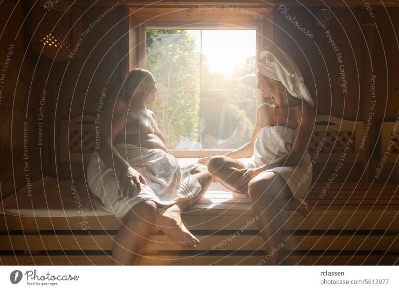 Man and woman sitting opposite each other in a finish sauna, both wrapped in towels, sunlit window at spa hotel sweat german finland finnish couple windows