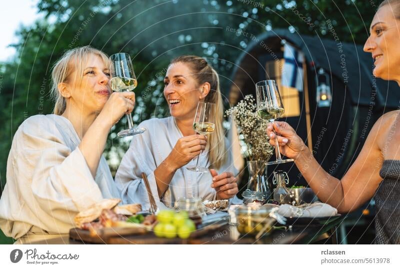 Women in bathrobes enjoying wine outdoors, with a  finnish mobile sauna cabin in the background women relaxation friends enjoyment leisure nature spa day