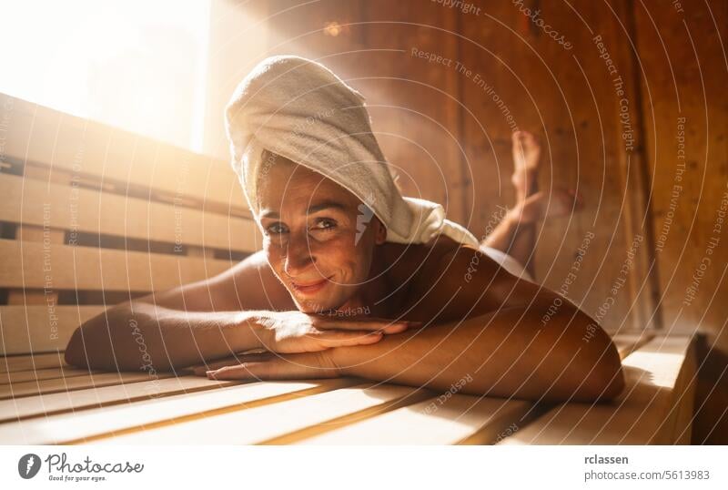 Woman relaxing in a finnish sauna, with a towel on her head, smiling at the camera bathrobe resort hotel woman relaxation wooden bench warmth wellness health