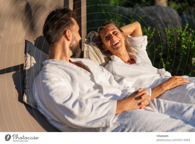 Couple in white bathrobes relaxing and laughing together on lounger in sunlight at a spa wellness hotel bench lounger resort love wellness spa couple