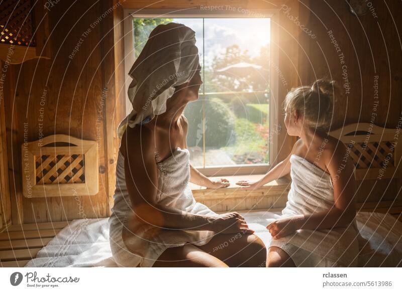 Back view of a mother and daughter in a finnish sauna, looking out a window, wrapped in towels at a spa wellness hotel windows bathrobe steam calm family woman