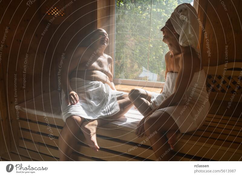Man and woman in a finnish sauna, laughing together, woman with a towel on her head, both seated at spa hotel wellness spa resort window sweat finland smiling