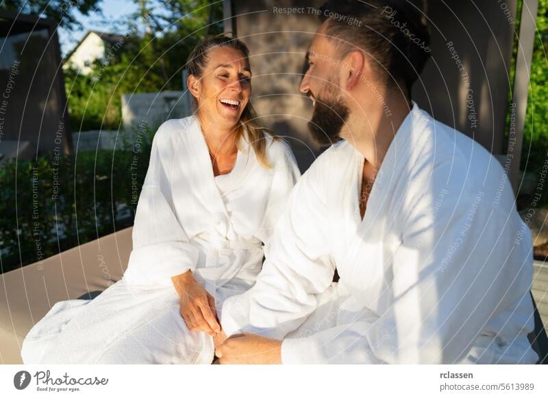 Two people in white robes sitting on a couple's lounger outdoors and having fun laughing together at a hotel love resort wellness spa relaxation smiling joy
