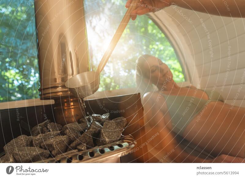 woman pouring water on sauna stones, another woman relaxing in the background. Mobile finish sauna concept image hot stones relaxation wellness bathing suits
