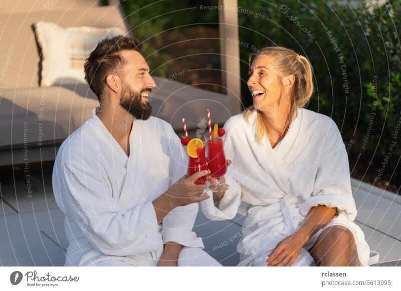 joyful couple in white bathrobes clinking glasses with red drinks, outdoors at golden hour at a hotel sipping wellness spa wellness resort white robes