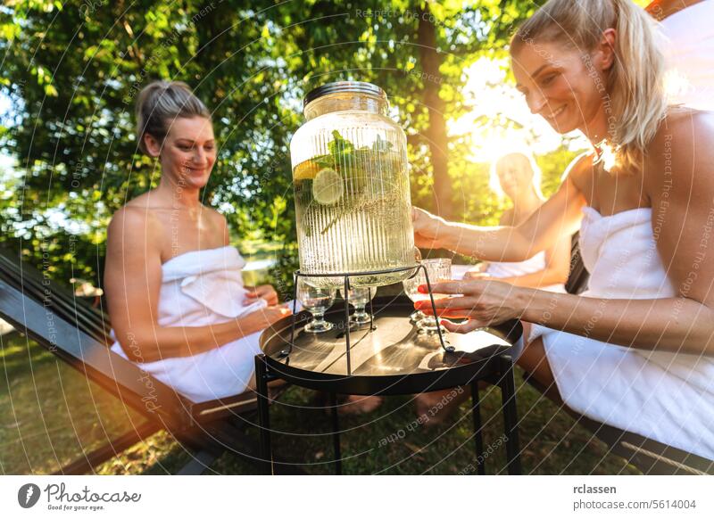 Women in towels serving and enjoying infused water from a dispenser with fresh lemons outdoors after sauna lime beverage dispenser women enjoyment hydration spa