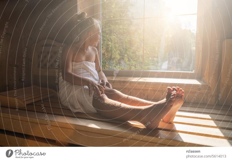 Child sitting on a finnish sauna bench, wrapped in a towel, looking out a sunlit window at a spa hotel bathrobe beauty spa calm comfortable enjoy sweat finland