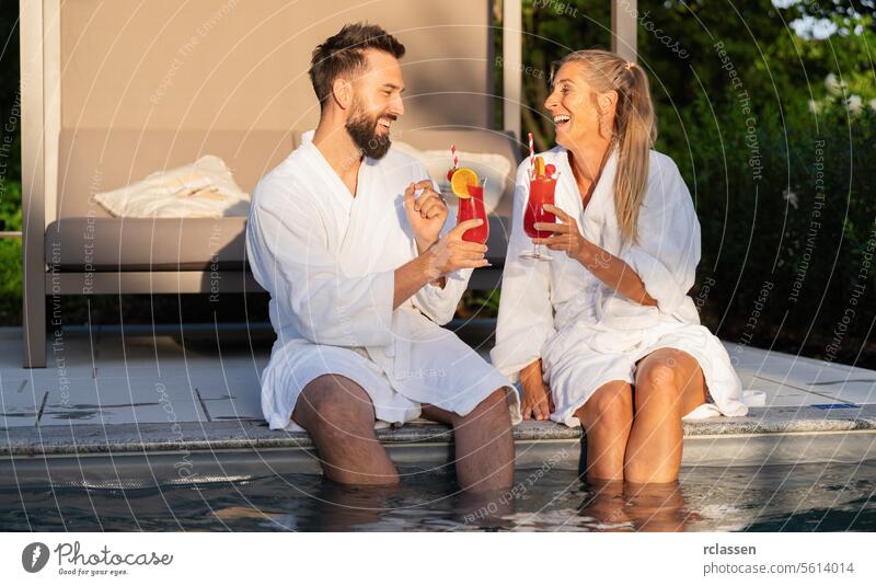 Two people in white bathrobes toasting with cocktails by the poolside, enjoying the sunset spa relaxation friends laughter clinking glasses wellness hotel