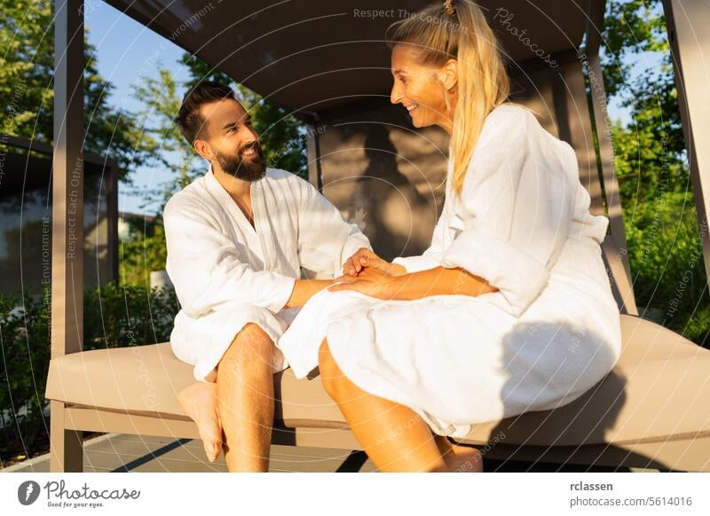Smiling couple in love in white bathrobes sitting on an outdoor bench lounger  in sunlight at spa wellness hotel smiling white robes relaxation happiness